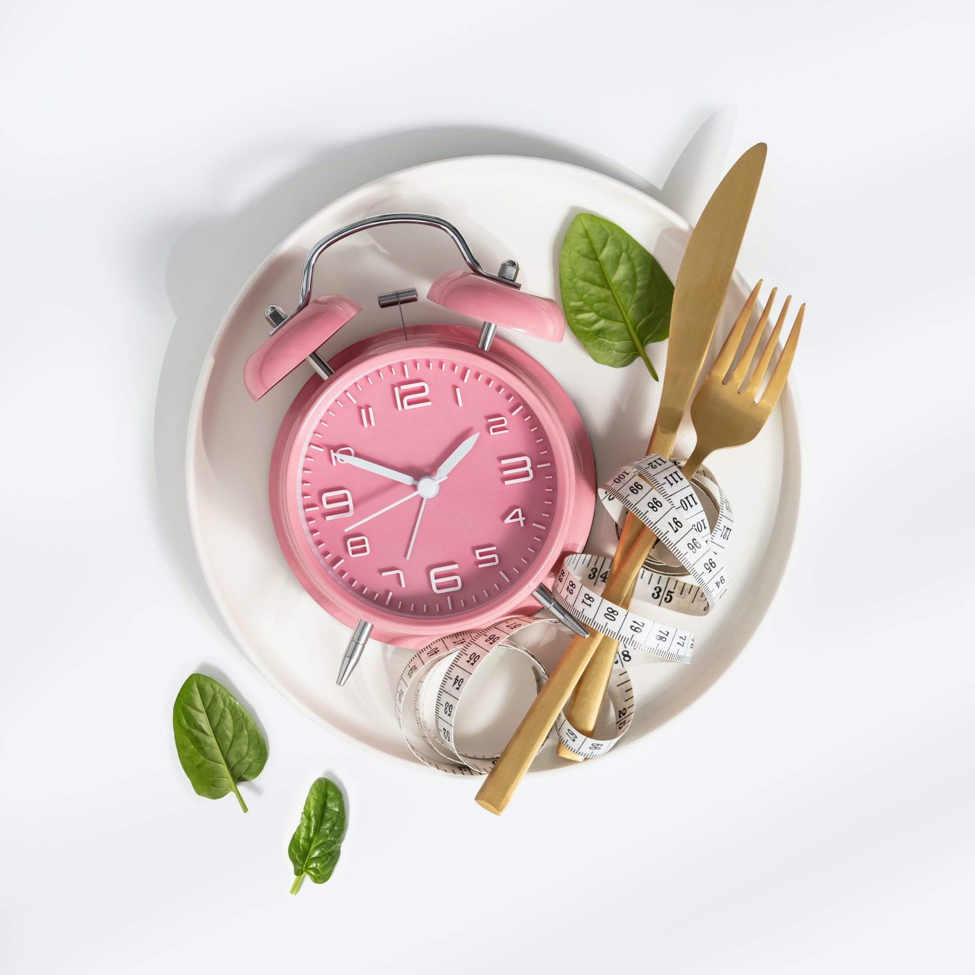 A pink alarm clock sits on a white plate with a measuring tape, gold fork, knife, and fresh spinach leaves, symbolizing healthy eating and time management.