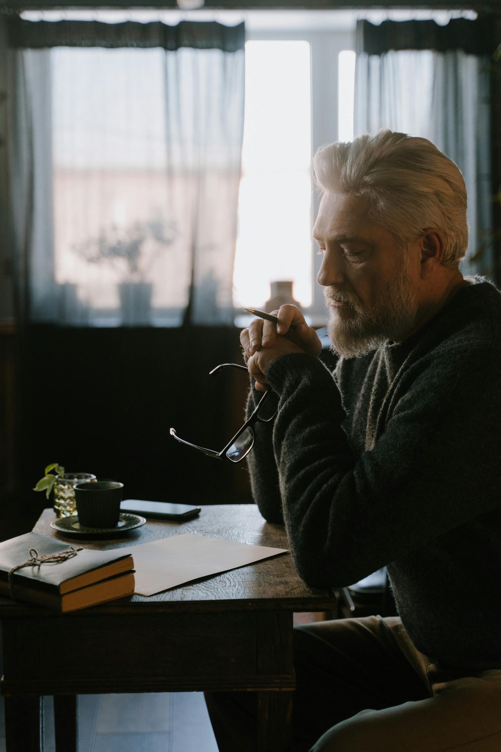 Pensive senior adult with white hair and glasses, writing at a wooden table indoors.