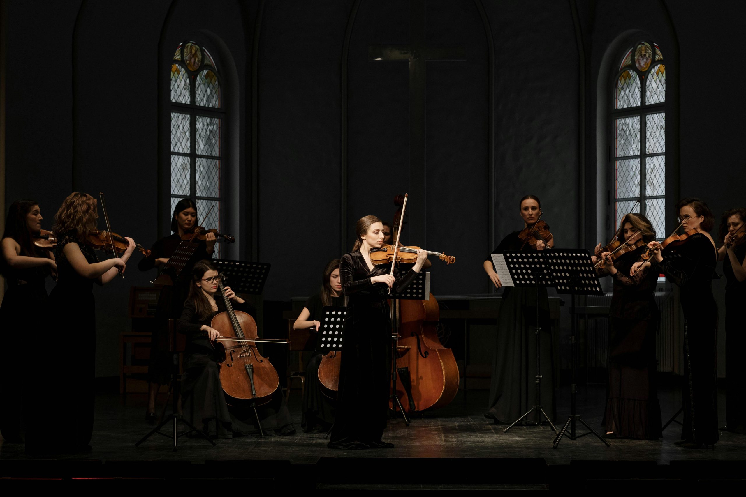 Women musicians perform with violins and cellos in a classical orchestra setting.